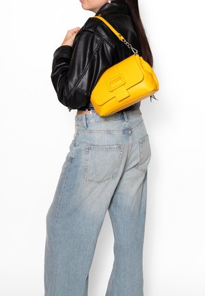 Femme portant un jean bleu clair et une veste en cuir noire, portant un sac à bandoulière jaune avec un rabat et un détail de boucle.