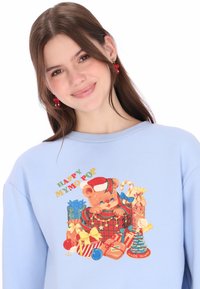 Sudadera azul claro con un gráfico de un oso de peluche con un gorro de Papá Noel, rodeado de regalos coloridos, bastones de caramelo y decoraciones navideñas.