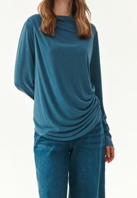 Sininen, pitkähiha blouse, jossa on laskostettu kaula-aukko ja kokoontunut sivuosa, valmistettu sileästä ja pehmeästä kankaasta. Yhdistetty sinisiin farkkuihin.