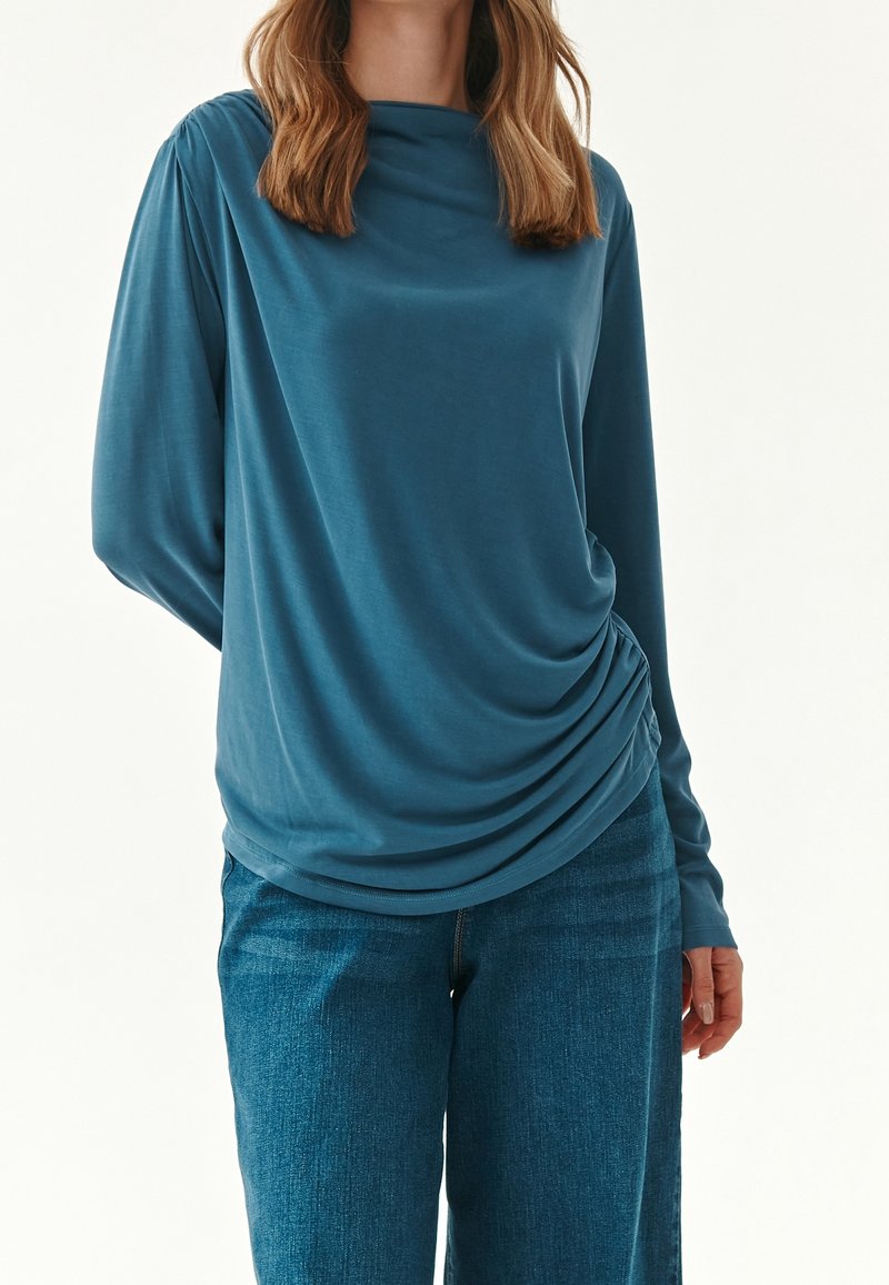 Sininen, pitkähiha blouse, jossa on laskostettu kaula-aukko ja kokoontunut sivuosa, valmistettu sileästä ja pehmeästä kankaasta. Yhdistetty sinisiin farkkuihin.