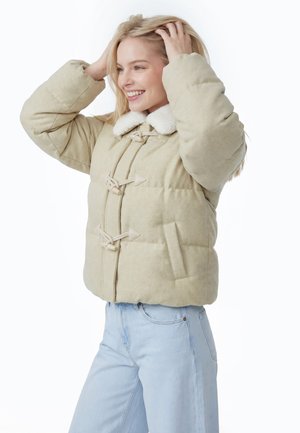 Smilende kvinde i lyse beige dynejakke med snoreknapper og hvid krave, der holder håret med begge hænder og har lyseblå jeans på.