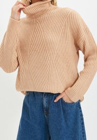 Jasnoróżowy sweter w prążki z golfem, z poziomymi i ukośnymi wzorami, o miękkiej strukturze i szerokich rękawach, zestawiony z niebieskimi dżinsami z wysokim stanem.