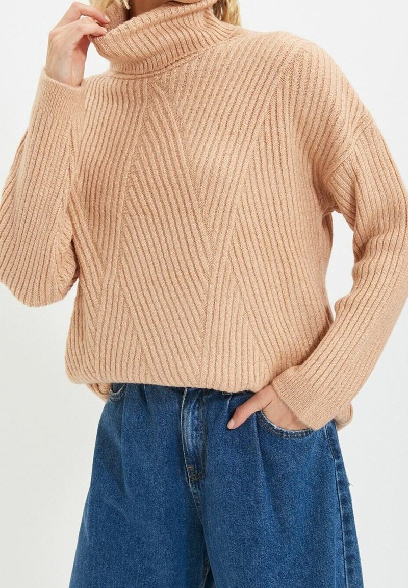 Jasnoróżowy sweter w prążki z golfem, z poziomymi i ukośnymi wzorami, o miękkiej strukturze i szerokich rękawach, zestawiony z niebieskimi dżinsami z wysokim stanem.