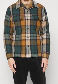 Muž ve zapnuté kárované bundě s náprsními kapsami v zelené, hořčicové, bílé a černé barvě, v kombinaci s černými kalhotami.