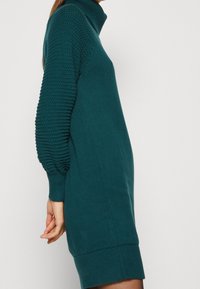 Robe tricotée teal avec un col montant et des manches bouffantes texturées ; présente des poignets et un ourlet côtelés, créant une silhouette ajustée.