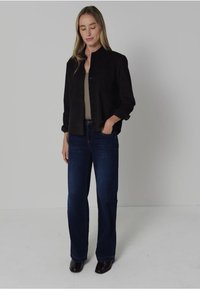 Vrouw met lang blond haar, gekleed in een zwarte button-up jas, een beige top, donkerblauwe jeans en zwarte schoenen, staand met één hand in haar broekzak.