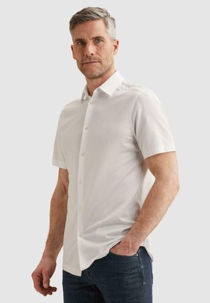 Midaldrende mand med gråt hår, iført en kortærmet hvid skjorte med knapper og mørkeblå jeans, stående med den ene hånd i lommen.