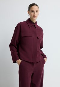 Burgundy oversized jas met een kraag, twee borstzakken en een centrale knoopsluiting, gecombineerd met bijpassende broek. Gladde textuur.