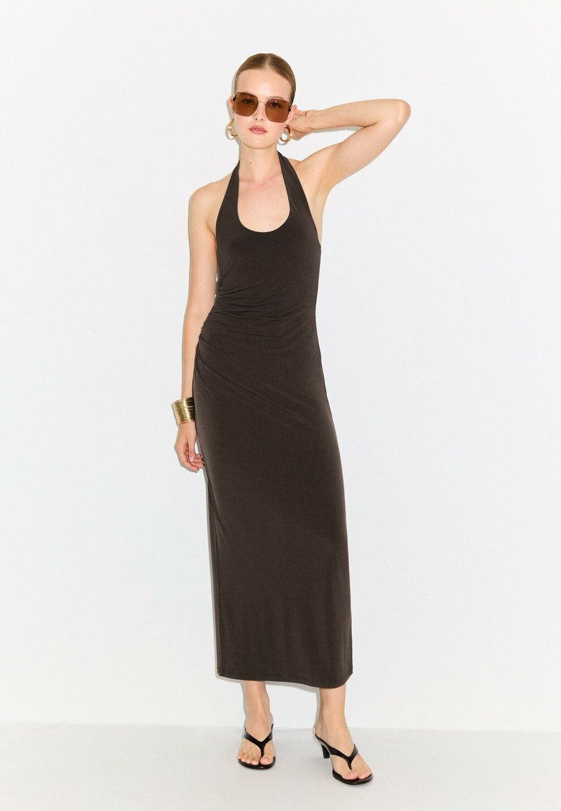 Femme portant une robe longue noire sans manches à col halter, des lunettes de soleil marron, des boucles d'oreilles créoles dorées, un bracelet manchette et des sandales à talons noires, posant.