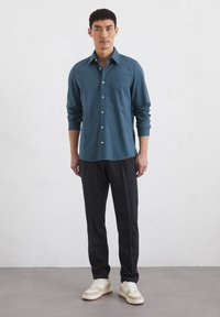 Teal geruit overhemd met lange mouwen en knopen, gecombineerd met zwarte broek en crème sneakers, staande tegen een witte textuur achtergrond.