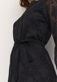 Svart spetsklänning med blommönster, långa ärmar och midjehögt bälte. Tyget är lätt och har en texturerad yta.