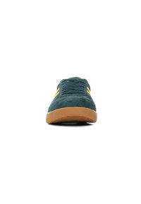 Zapatillas de ante verde con suelas de goma, que presentan cordones planos y rayas amarillas en el costado. Punta redondeada y diseño de caña baja.