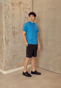 Jeune homme en t-shirt bleu, shorts noirs et baskets noires, appuyé contre un mur d'angle beige texturé, les mains dans les poches.
