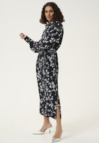 Robe midi florale en navy avec des fleurs blanches, présentant un devant à boutons, des manches longues, une taille à nouer et des fentes sur les côtés. Des talons argentés complètent le look.