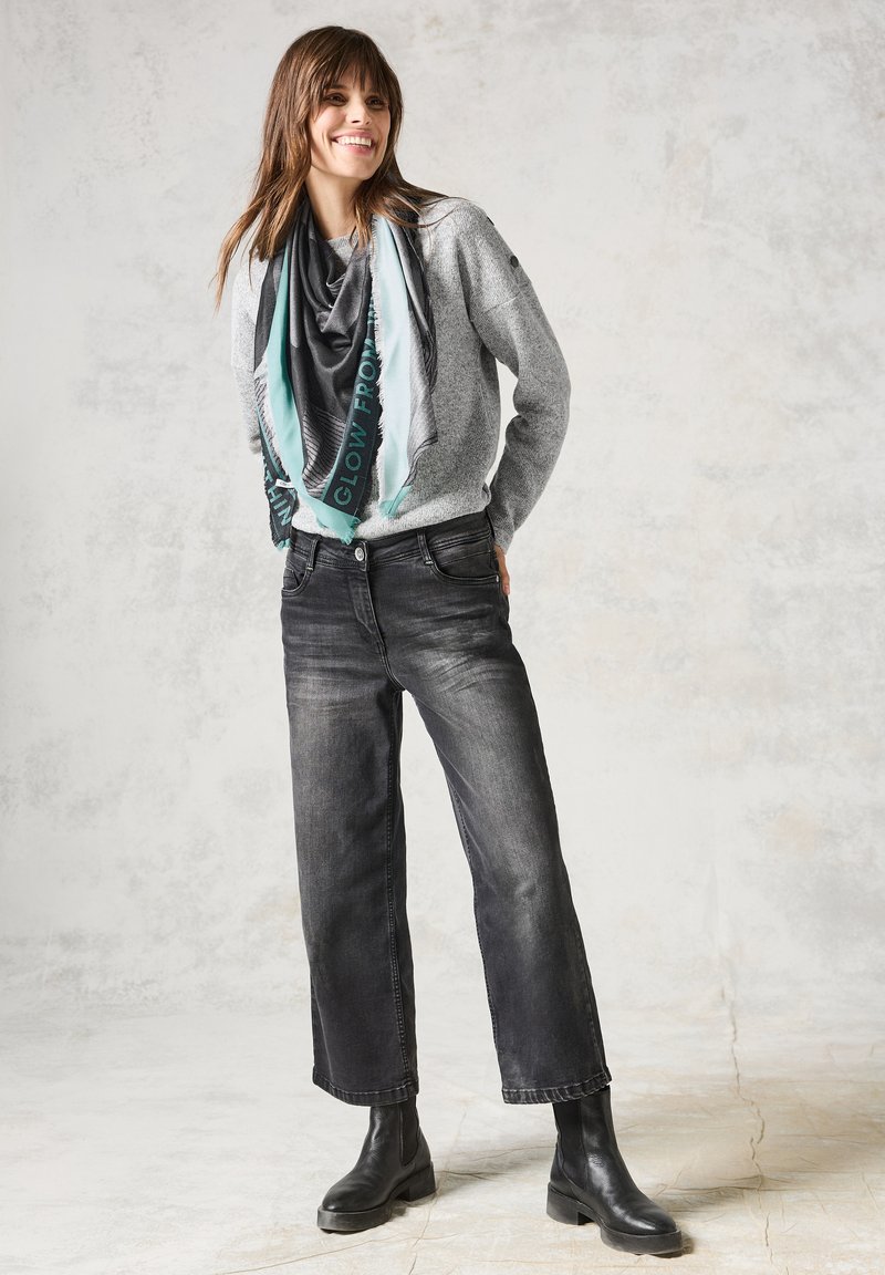 Suéter gris de punto, bufanda gris y verde azulado con estampado, jeans negros de pierna ancha y botines negros. Texturas suaves, ajuste casual, diseño en capas.
