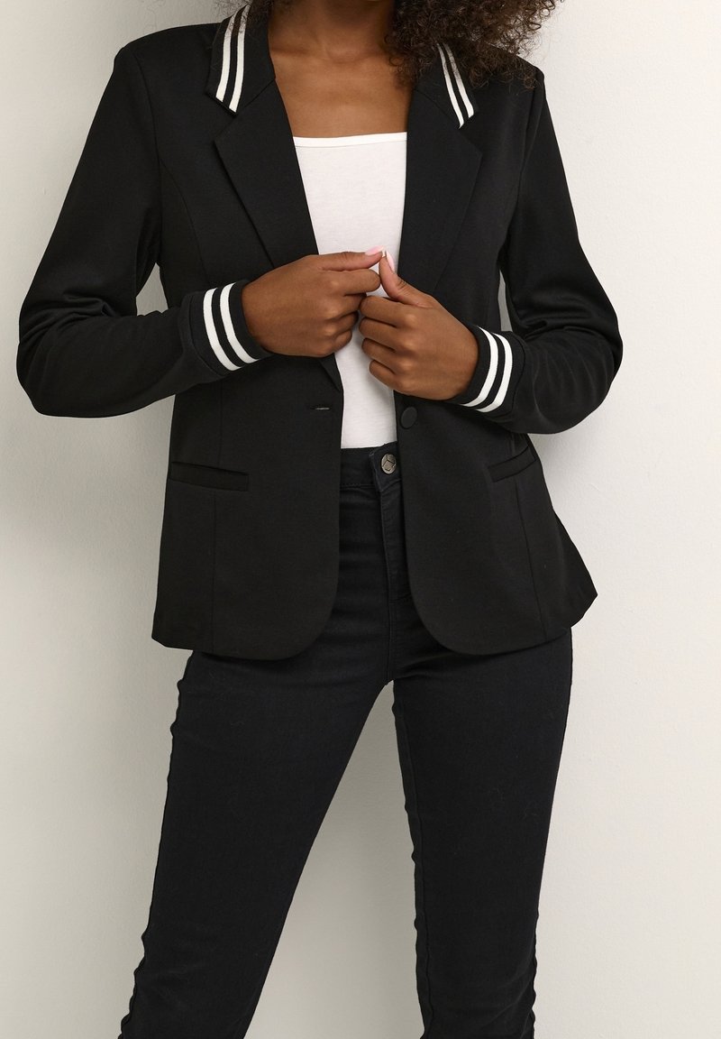 Musta bleiseri, jossa valkoiset raidalliset yksityiskohdat kauluksessa ja hihansuiden kohdalla. Yhdellä napilla suljettava ja kaksi etutaskua.