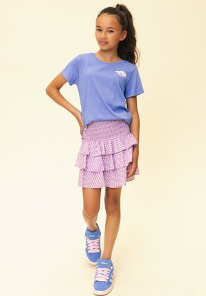 Meisje met lang haar in een paardenstaart draagt een blauw T-shirt, een roze geplooide rok met patroon en blauwe Adidas sneakers, staand met één hand op de heup.