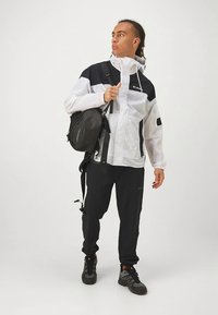 Valge ja must kerge jope kõrge kraega, millele on kombineeritud mustad püksid ja kingad. Komplektis on must seljakott reguleeritavate rihmadega.