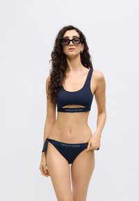 Σετ μπικίνι σε ναυτικό μπλε χρώμα με τοπ σε σχήμα σούπερ-νέκ με λεπτομέρεια κοψίματος και ψηλόμεσα εσώρουχα δεμένα στα πλάγια. Υφή υφάσματος με λογότυπο.