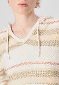 Hættebluse i off-white med vandrette striber i fersken, guld og beige. Har en tekstureret strik og snøre detalje.