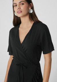 Schwarzes Wickelkleid mit kurzen Ärmeln, ausgestattet mit einem V-Ausschnitt und einer seitlichen Taillierung. Der Stoff ist leicht, hat eine glatte Textur und eine subtile Drapierung.