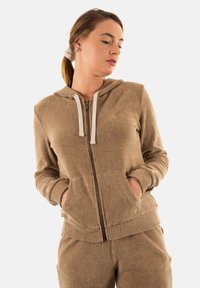 Sudadera con cremallera de color marrón y con superficie acanalada, que presenta una capucha con cordón, bolsillos frontales y puños ajustados. Se combina con pantalones a juego.