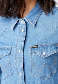 Light blue denim shirt featuring a pointed collar, two chest pockets with white snaps, and zigzag stitching on the pocket flaps. Wrangler logo visible.