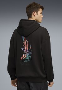 Jonge man in een zwarte hoodie met een kleurrijk abstract neonontwerp op de rug, staand tegen een effen grijze achtergrond, met de zijkant van zijn gezicht naar voren. 