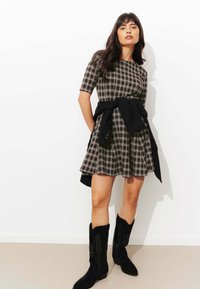 Robe à carreaux dans des nuances de gris et de marron, à manches courtes, au design évasé, associée à un pull noir noué à la taille, et des bottes de cow-boy noires.