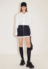 Witte overhemd met knopen, zwarte mini rok met grote zakken en rits, kniehoge zwarte sokken, stevige zwarte sneakers en een zwarte pet.