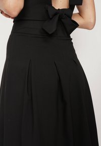 Czarna sukienka z dopasowaną górą, plisowanym dołem i akcentem w postaci kokardy w talii. Gładki, lekko teksturowany materiał.