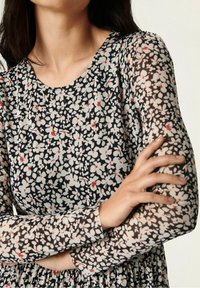 Vestido con estampado floral de base negra, con flores blancas y rojas. Mangas largas y transparentes, y un escote redondeado. Textura de tela suave.