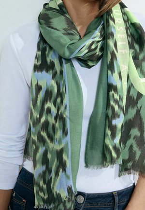 Écharpe à motifs verts, noirs et bleus avec bords effilochés portée sur une chemise blanche à manches longues et un jean bleu foncé.