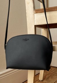 Musta nahkainen olkalaukku pyöreällä ylhäällä, vetoketjukiinnityksellä ja kohokuvioidulla logolla edessä. Laukussa on ohut hihna ja sileä pinta.