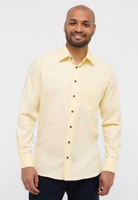 Camicia a maniche lunghe di un leggero giallo con colletto a punte, dotata di taschino sul petto e dettagli con bottoni scuri. Tessuto morbido, vestibilità sagomata.