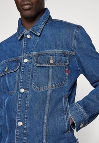 Denim jas in diepblauw met een klassieke snit, voorzien van twee voorzakken, knoopsluitingen en stikselaccenten. Gladde textuur.