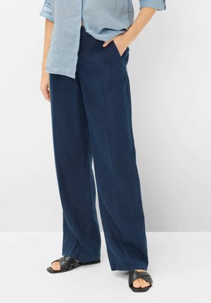 Persoon draagt losse donkerblauwe broek, lichtblauw opgerolde mouwen overhemd en zwarte open teen sandalen, staat met één hand in zak.