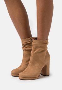 Bottes cheville en daim beige avec un design souple, un talon bloc haut, une fermeture éclair latérale, une texture lisse et un bout arrondi.