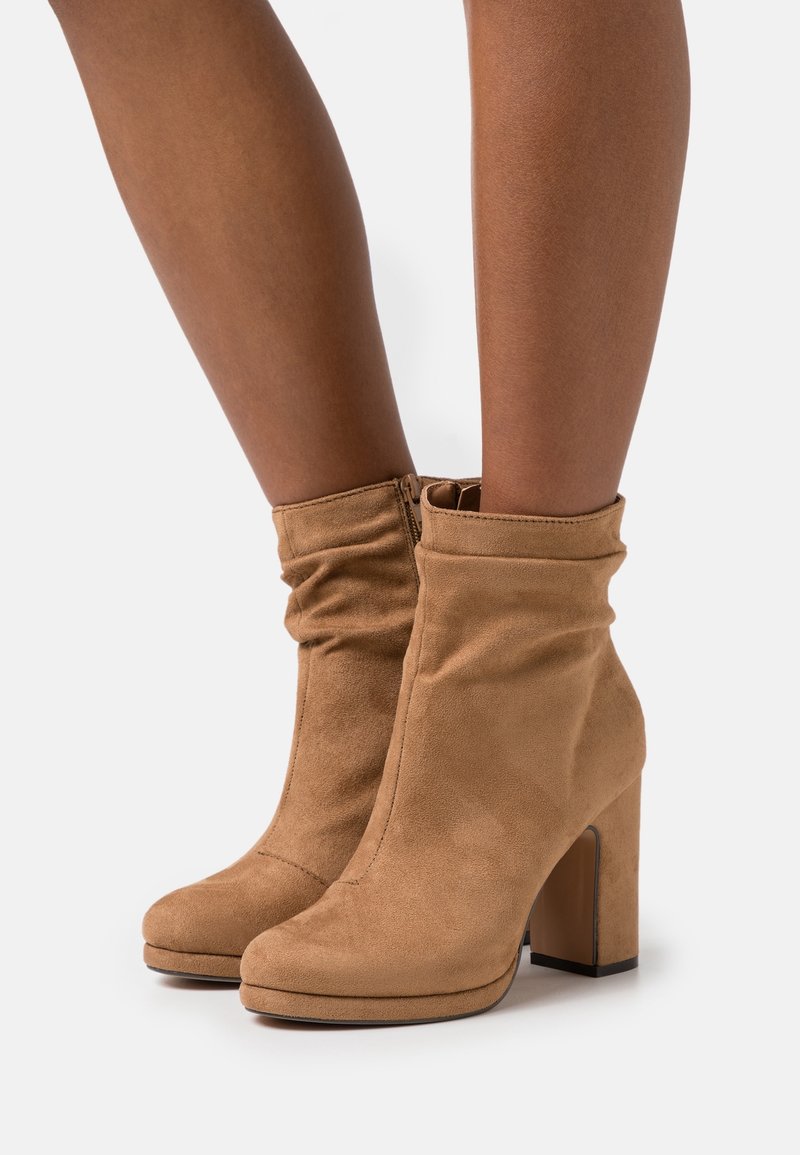 Bottes cheville en daim beige avec un design souple, un talon bloc haut, une fermeture éclair latérale, une texture lisse et un bout arrondi.