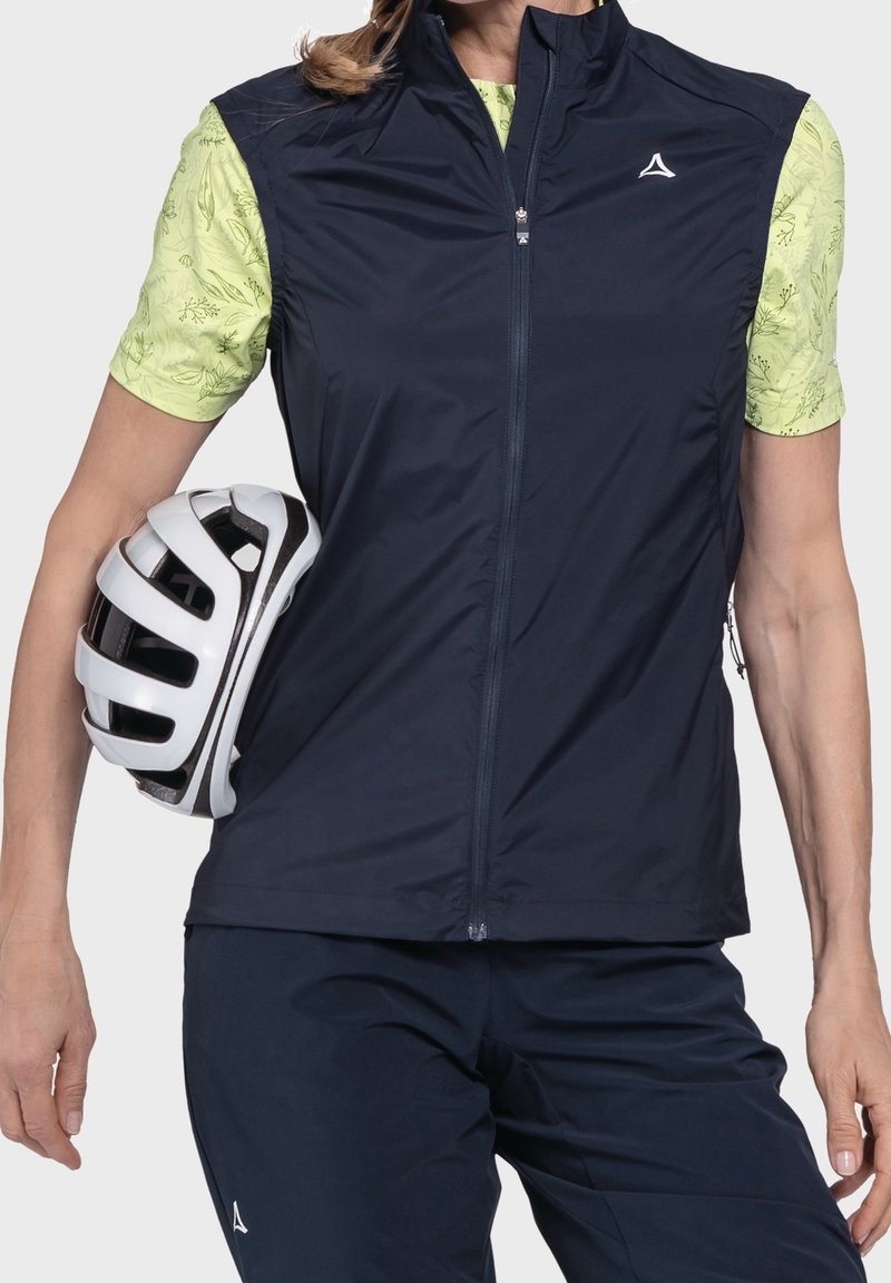 Personne portant un gilet de cyclisme bleu marine et un short, tenant un casque de vélo blanc et noir contre son côté, portant un t-shirt vert clair à manches courtes avec des motifs de feuilles.