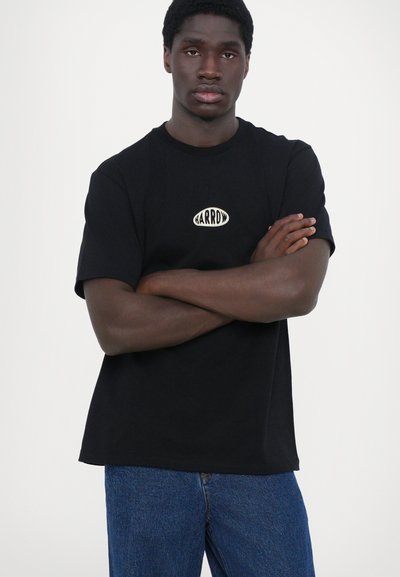 Camiseta negra de manga corta con un logo circular que dice "BARROD" en crema, combinada con jeans de mezclilla. Textura suave, corte relajado.