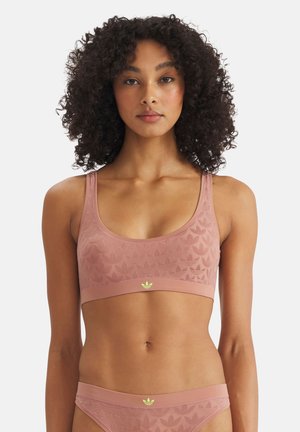 Nainen, jolla on kiharat hiukset ja joka pukeutuu vaaleanpunaisen sävyiseen, yhteensopivaan urheilurintaliiviin ja alushousuihin, joissa on hienovaraiset kohokuvioidut logot valkoisella taustalla.