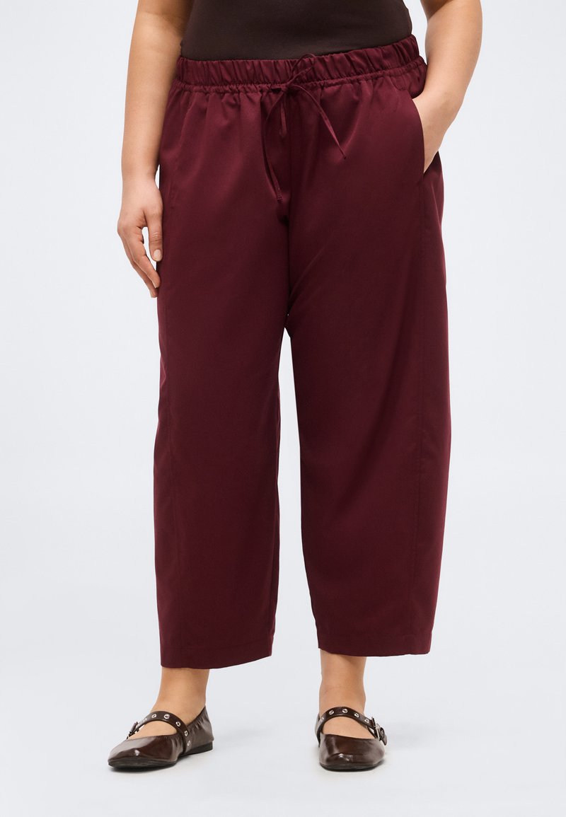 Άτομο που φορά χαλαρό, κοντό μπορντό παντελόνι με κορδόνι στη μέση και καφέ επίπεδα παπούτσια με λουράκια και καρφιά.