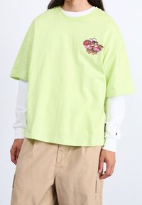 Limegrünes Baumwoll-T-Shirt mit kurzen Ärmeln, das ein pinkes und braunes grafisches Design auf der linken Brust aufweist. Darunter wird ein weißes Langarmshirt getragen.