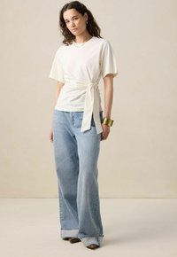 Haut beige clair à manches courtes avec un détail noué à la taille, associé à un jean en denim bleu clair à jambes larges et un bracelet en métal doré.