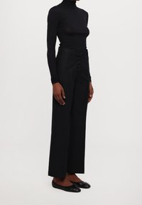 Svart, långärmad polotröja i kombination med högmidjade svarta byxor med sex knappar. Båda är figurnära och har en texturerad yta.