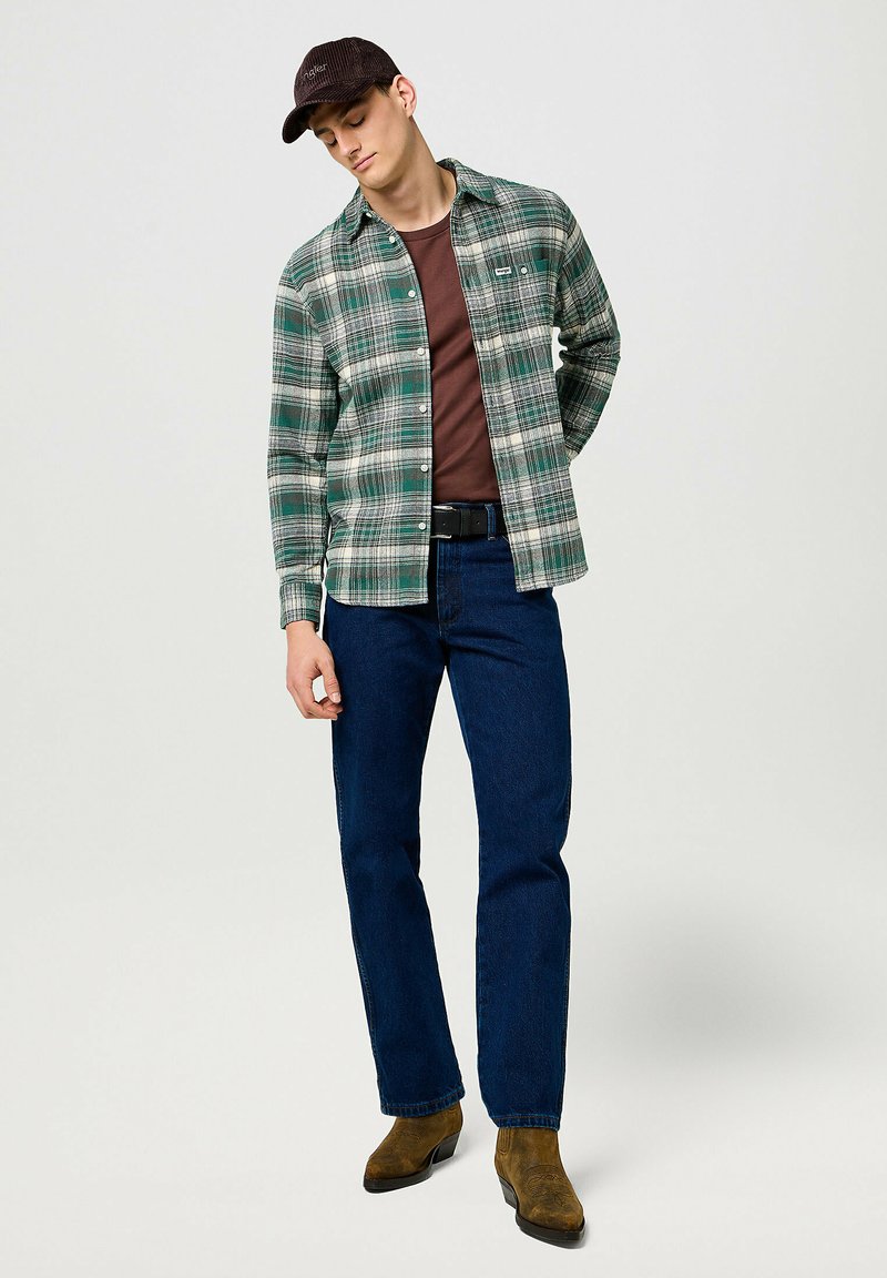 Grünes und weißes karierter Flanellhemd, über einem braunen Hemd getragen, kombiniert mit dunkelblauen Jeans und braunen Cowboy-Stiefeln. Schwarze Kappe getragen.