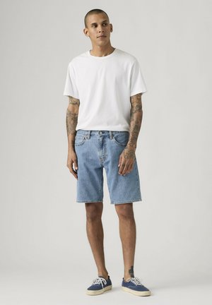 Joven tatuado que lleva una camiseta blanca, pantalones cortos de mezclilla azul claro y zapatillas azul marino, de pie contra un fondo claro y liso.