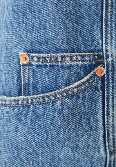Tejido de mezclilla azul con un bolsillo frontal que presenta costura naranja y un detalle de remache de cobre, mostrando fibras texturizadas en un corte clásico.