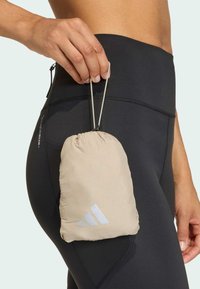 Beige, pakket trekkpose laget av lett materiale, med en hvit logo på forsiden. Holdt i en hånd ved siden av svarte leggings.